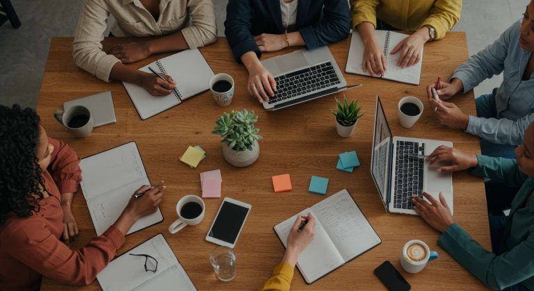 קבוצת נשים יזמיות ואמהות משתפות פעולה בסביבה חמה ומעוררת השראה, עם מחשבים וניירות עבודה, מסמלת קהילה ותמיכה עסקית.